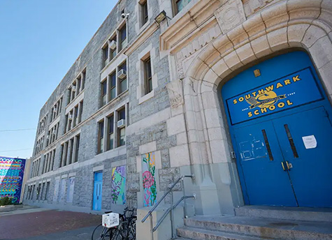 A four-story stone building with several windows and a staircase leading up to the blue front door that reads “Southwark School” above.