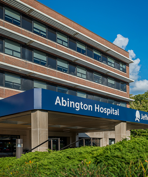 The exterior of a multi-story, brick building with the words “Abington Hospital - Jefferson Health” on a blue backdrop above the entranceway with green shrubbery in front of it.