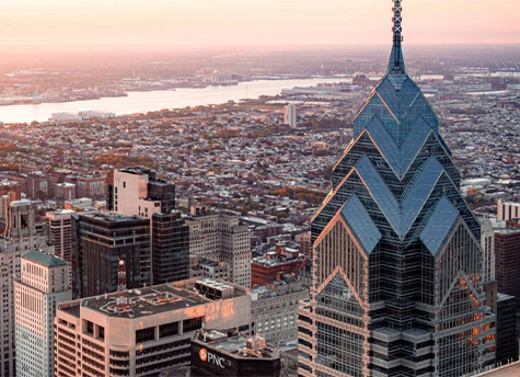 An aerial shot of the Philadelphia skyline with the river and a yellow-pinkish sky in the background and several tall buildings in the foreground.