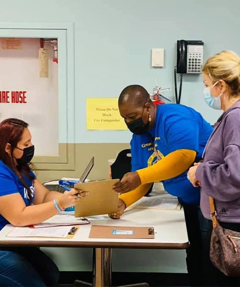 An ACLAMO volunteer wearing a mask sits at a table while looking over a clipboard with another volunteer in a mask who is standing while a visitor also wears and watches the volunteers.