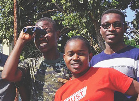 A group of participants stand in front of a building and some trees while one of them uses a pair of binoculars, one holds a drone, and most smile at the camera.