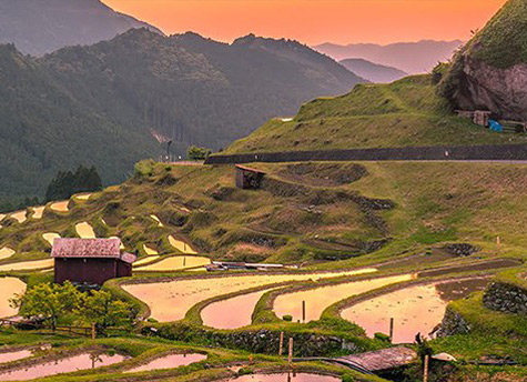 Some small structures sit amongst several small ponds along a hillside overlooking lush green mountains with an orange sky in the background.