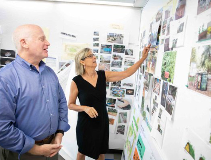 Drew Harris and Kim Douglas stand in a white room covered in printed out images and graphics while looking at one of the images together.