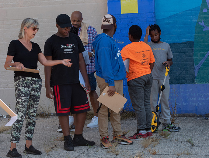 Kim Douglas holds a clipboard in one hand and has her other arm outstretched as she speaks to a Park in a Truck worker while standing in an empty lot with other workers in the background.
