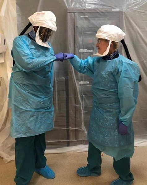 Two medical workers wearing full personal protective equipment share a fist bump while smiling at each other.