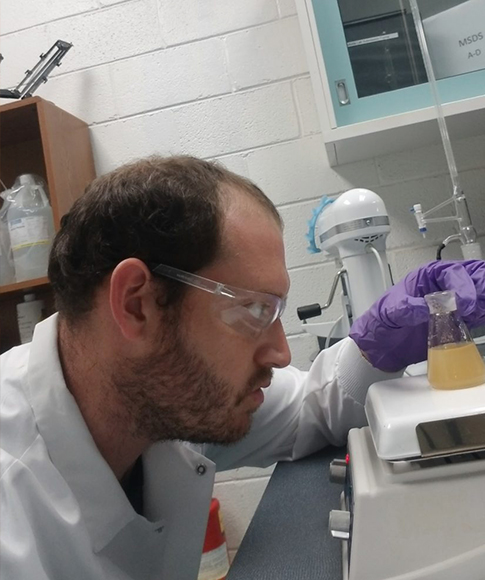 Seth Michael Martin wears a lab coat, safety glasses and gloves while examining a liquid sample in a lab.
